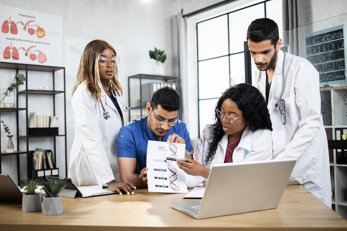 Group of four multiethnic diverse doctors genetics, having online meeting with colleague on laptop indoors, explaining DNA structure and discussing molecular formulas and genetic desease